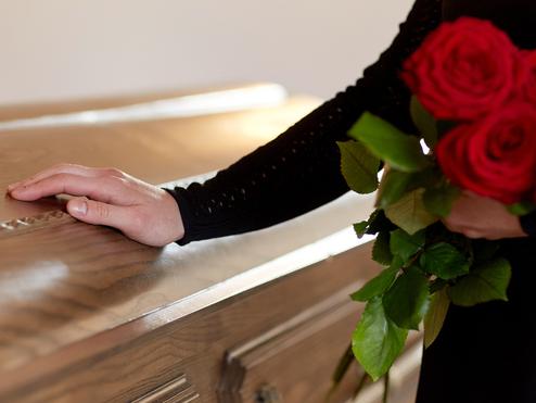 Eine Hand mit einer roten Rose berührt den Deckel eines holzvertäfelten Sargs in einem sanften Licht.