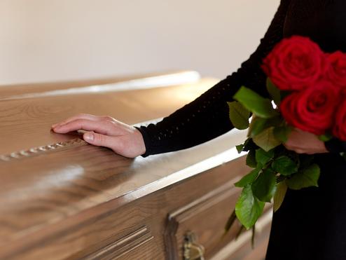 Eine Hand mit einer roten Rose berührt den Deckel eines holzvertäfelten Sargs in einem sanften Licht.