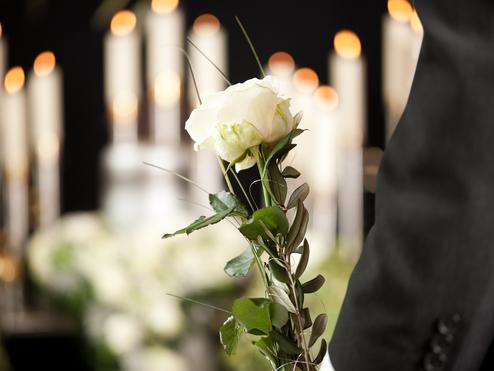 Weiße Rose mit Blättern in der Hand eines Mannes, im Hintergrund brennende Kerzen.