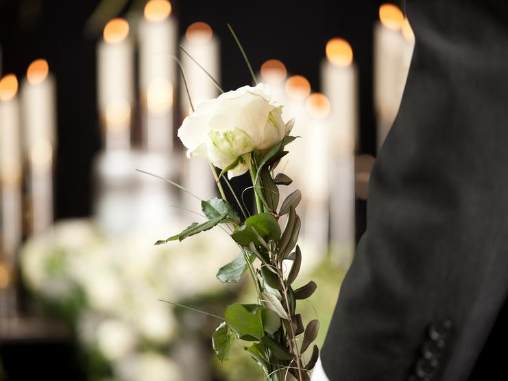 Weiße Rose mit Blättern in der Hand eines Mannes, im Hintergrund brennende Kerzen.