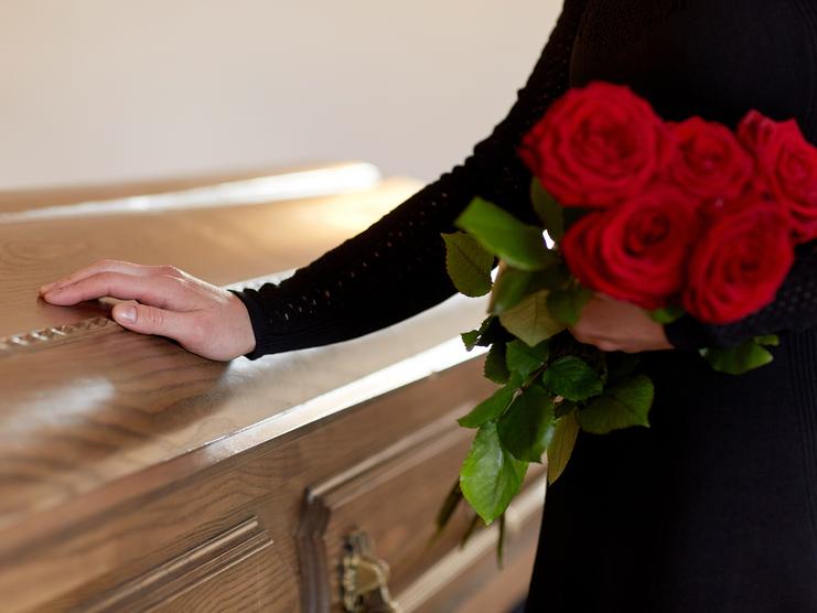 Eine Hand mit einer roten Rose berührt den Deckel eines holzvertäfelten Sargs in einem sanften Licht.