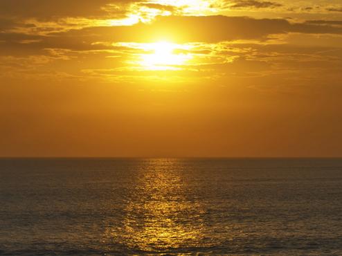 Sonnenuntergang über dem Meer, reflektiert goldenes Licht auf der Wasseroberfläche und dunklen Felsen im Vordergrund.
